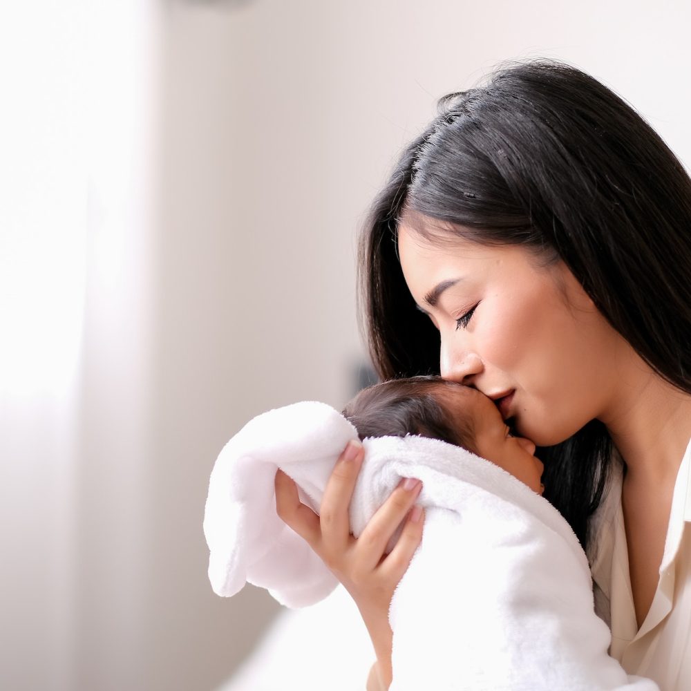 White shirt Asian mother is kissing at forehead of her newborn baby in bedroom