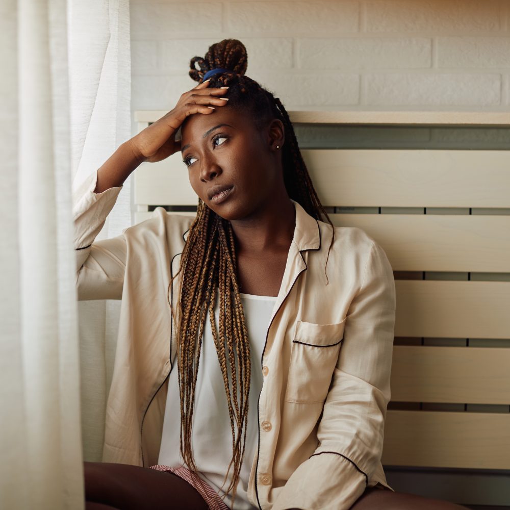 Young sad African American woman looking through the window at home.